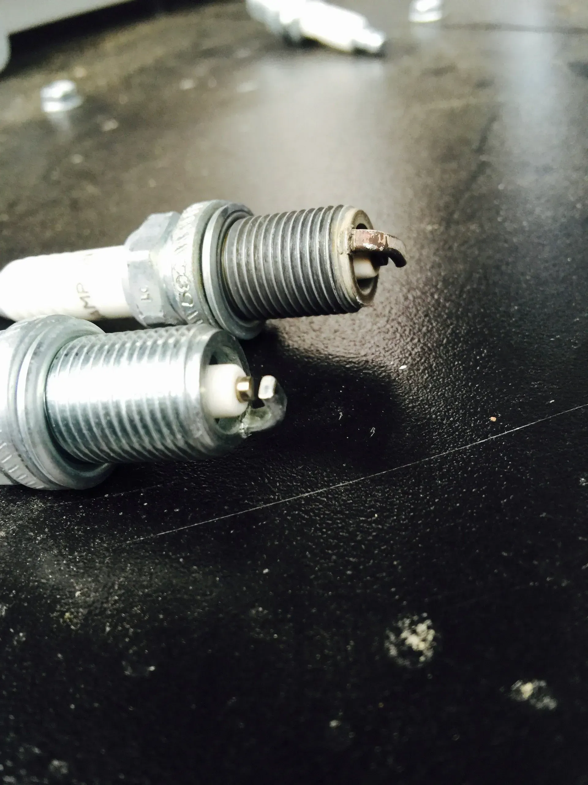 Two used spark plugs on a dark surface, one with a damaged tip.