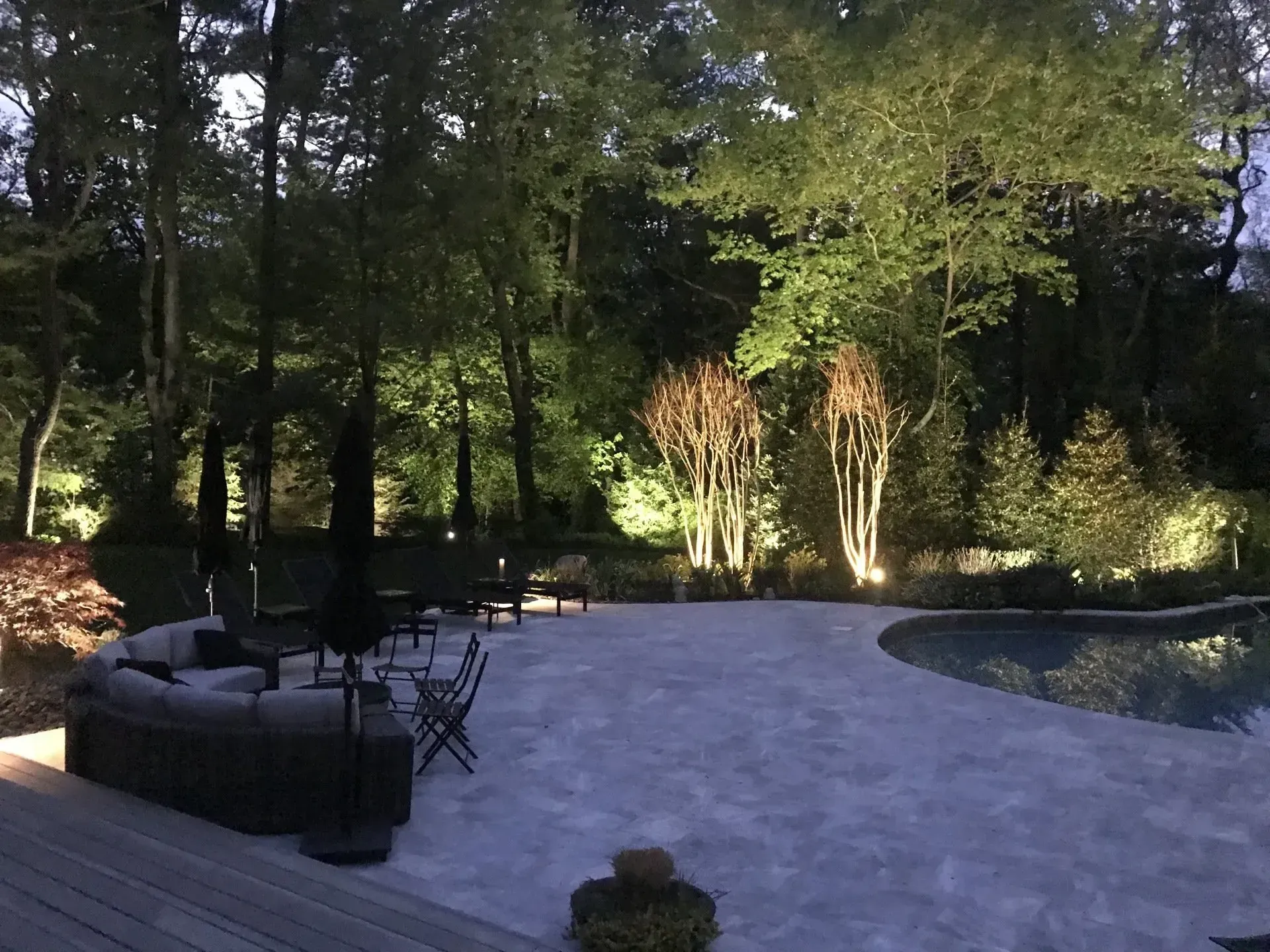 Night scene: lit patio with outdoor furniture, pool, trees.