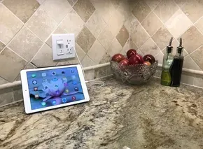 IPad on kitchen counter, next to outlet, bowl of apples, and oil bottles. Tile backsplash.