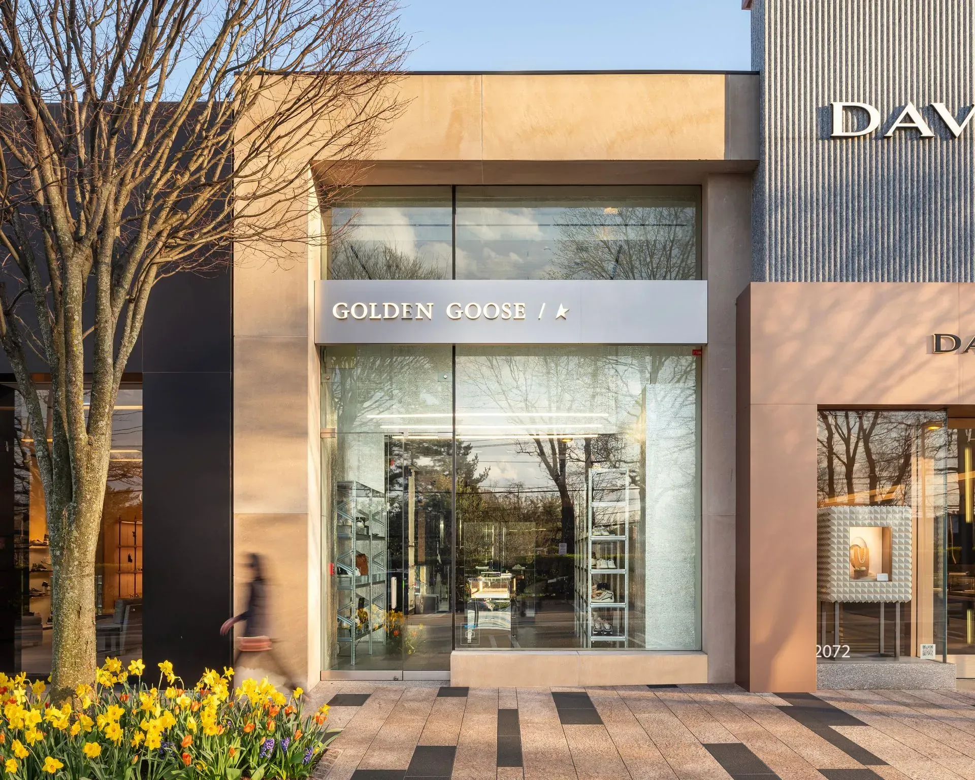 Golden Goose store with glass doors, silver sign. Person walks past. Yellow flowers in the foreground.