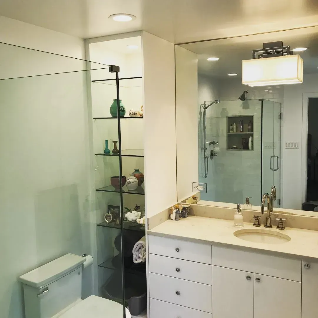Bathroom with white vanity, mirror, shower, and glass-door shelving filled with objects. Toilet in the corner.