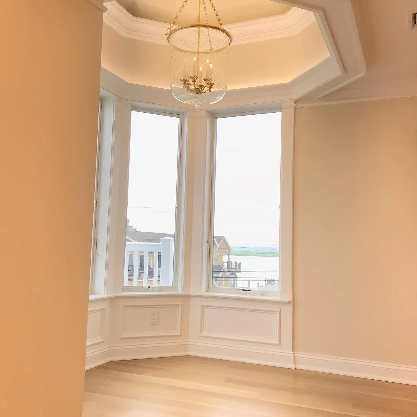 Empty room with large bay windows, wood floors, and a chandelier. Light beige walls and trim.