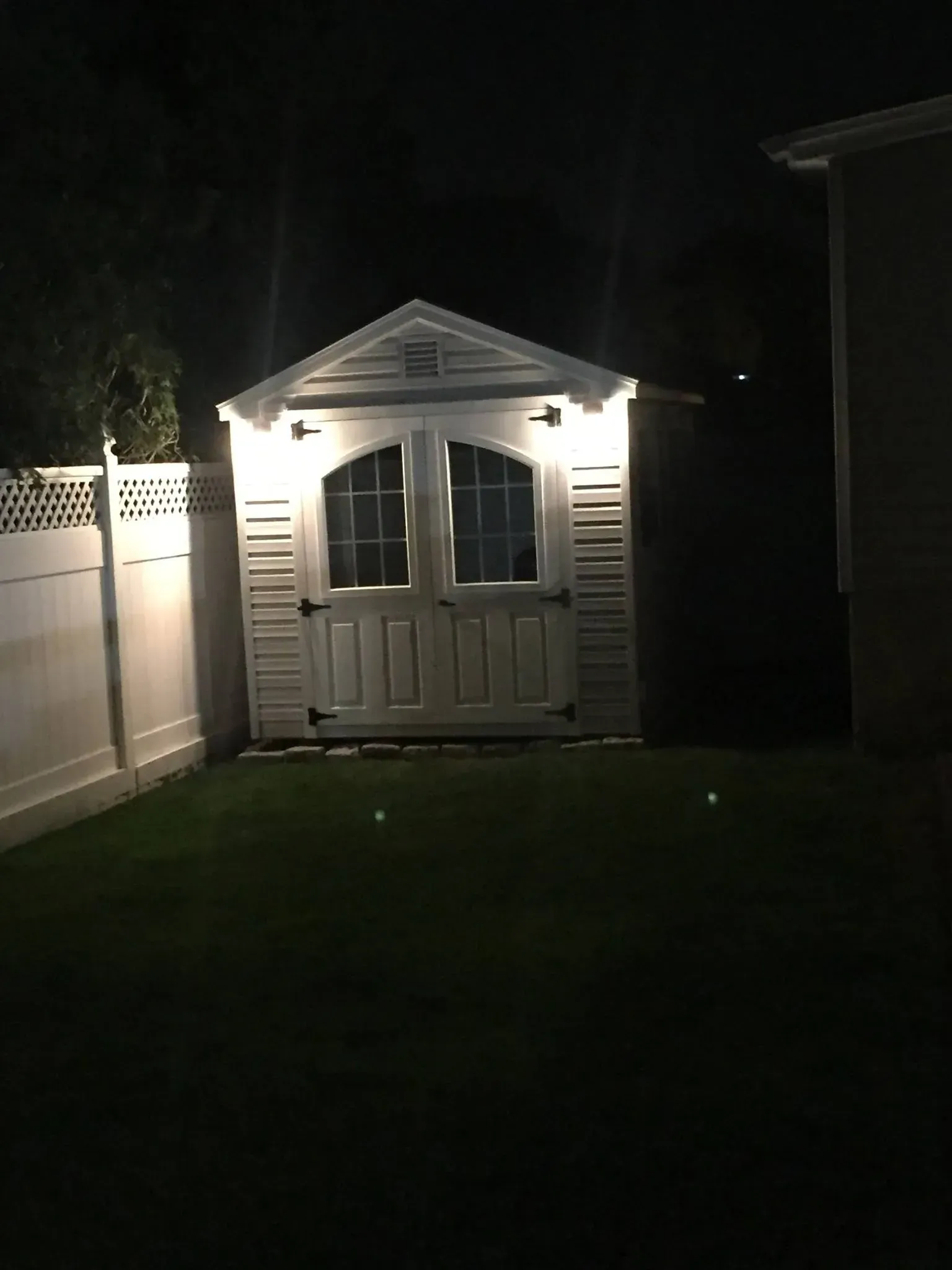A shed illuminated by lights at night, standing in a backyard, beside a white fence.