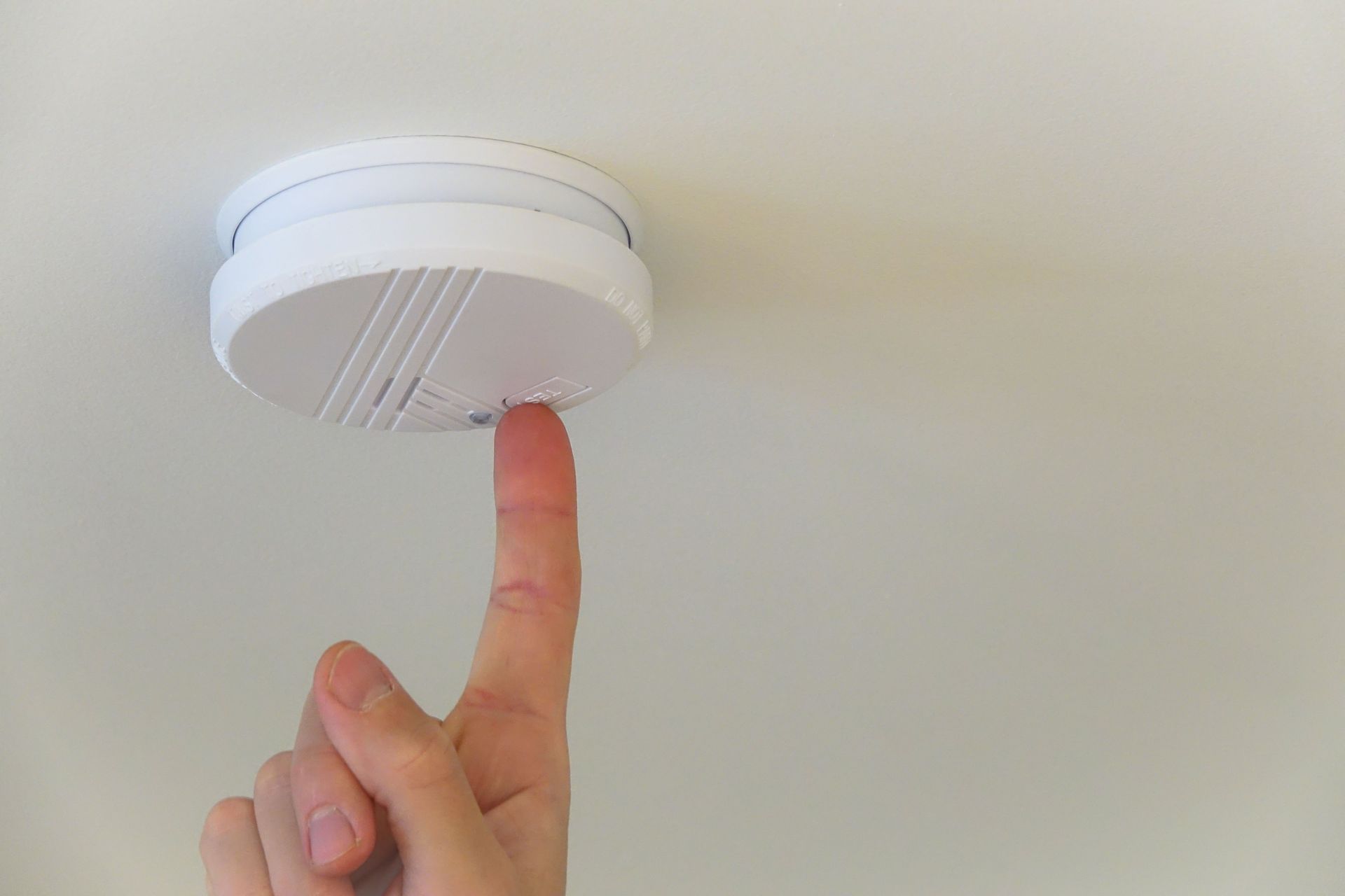 Finger pressing a button on a white smoke detector attached to a white ceiling.