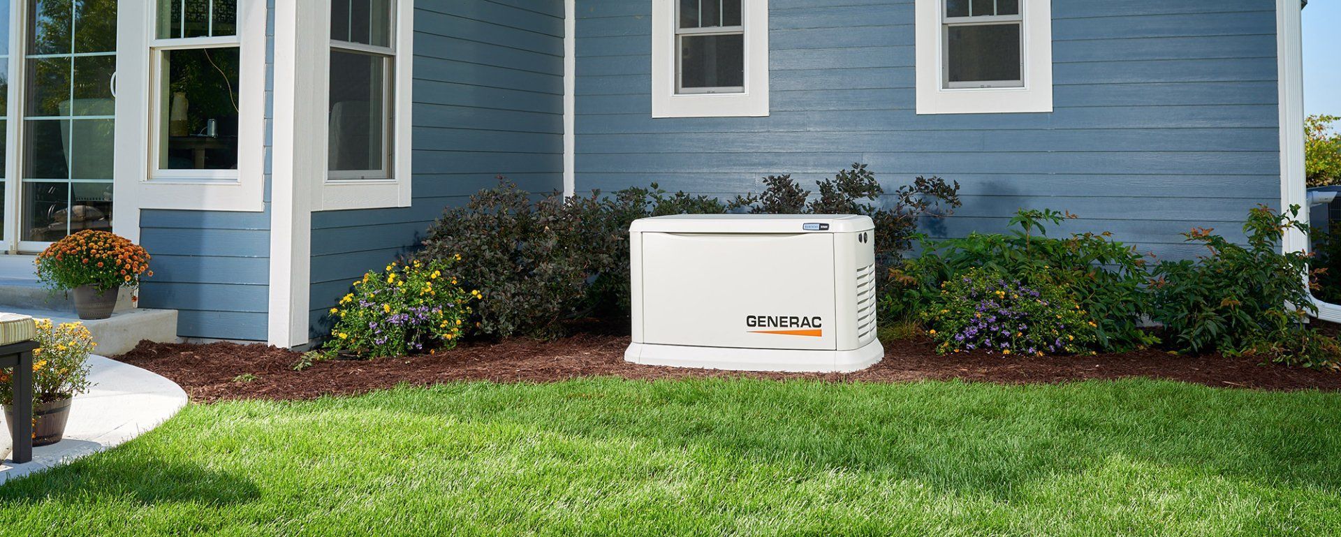 A white home generator is installed on the side of a blue house with green grass.