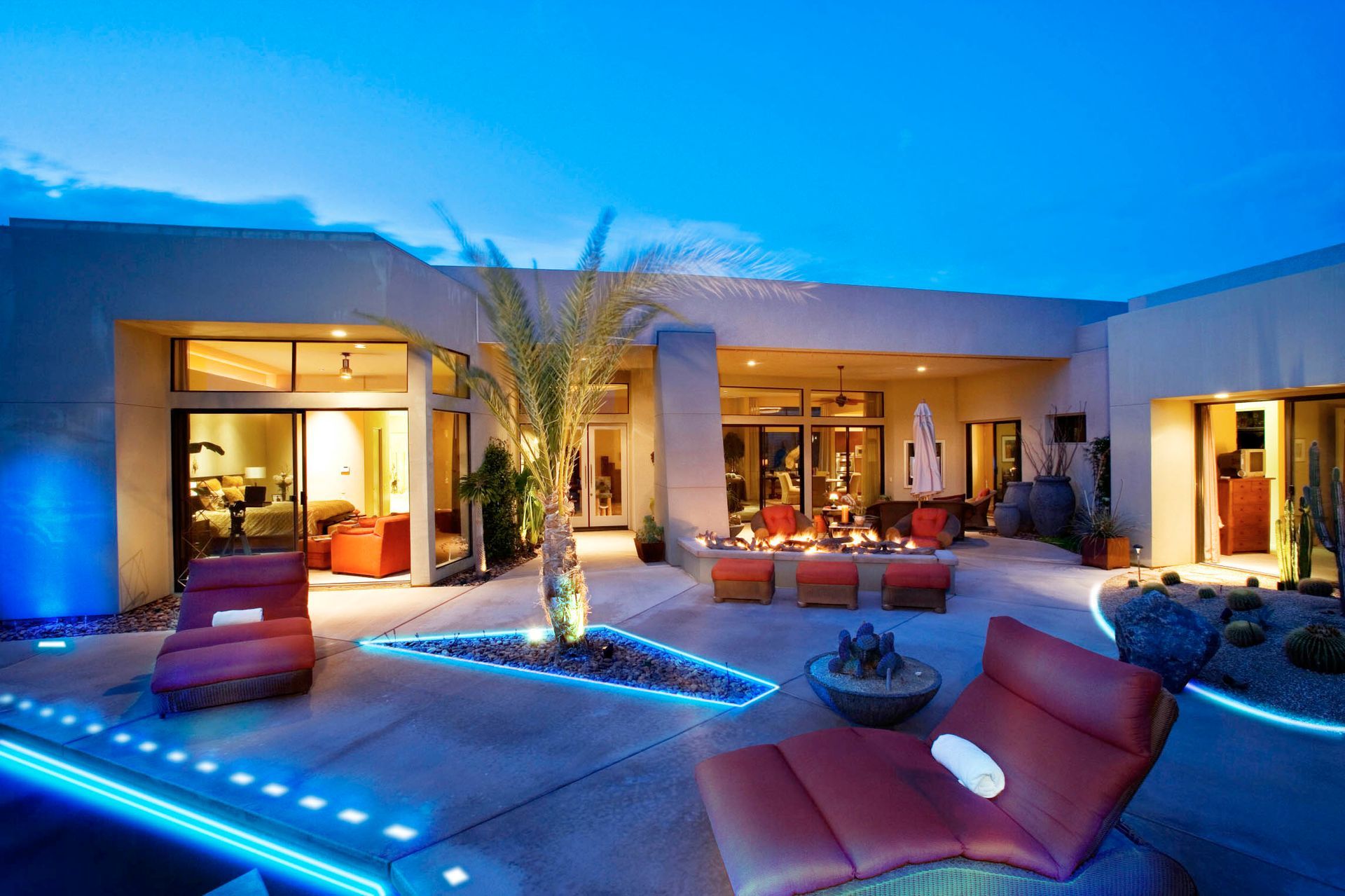 Patio with lounge chairs, fire pit, and palm tree, illuminated by blue lights at dusk.
