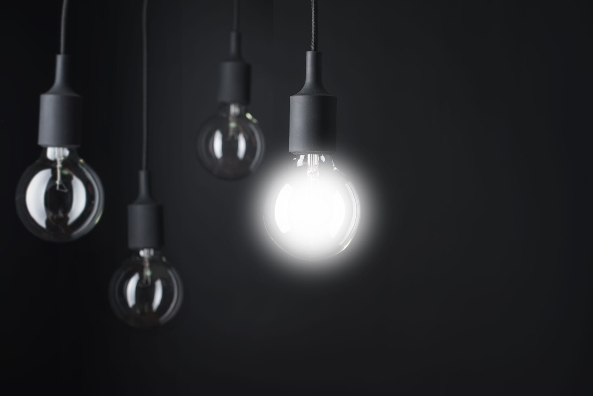 Four hanging lightbulbs, one illuminated, against a dark background.