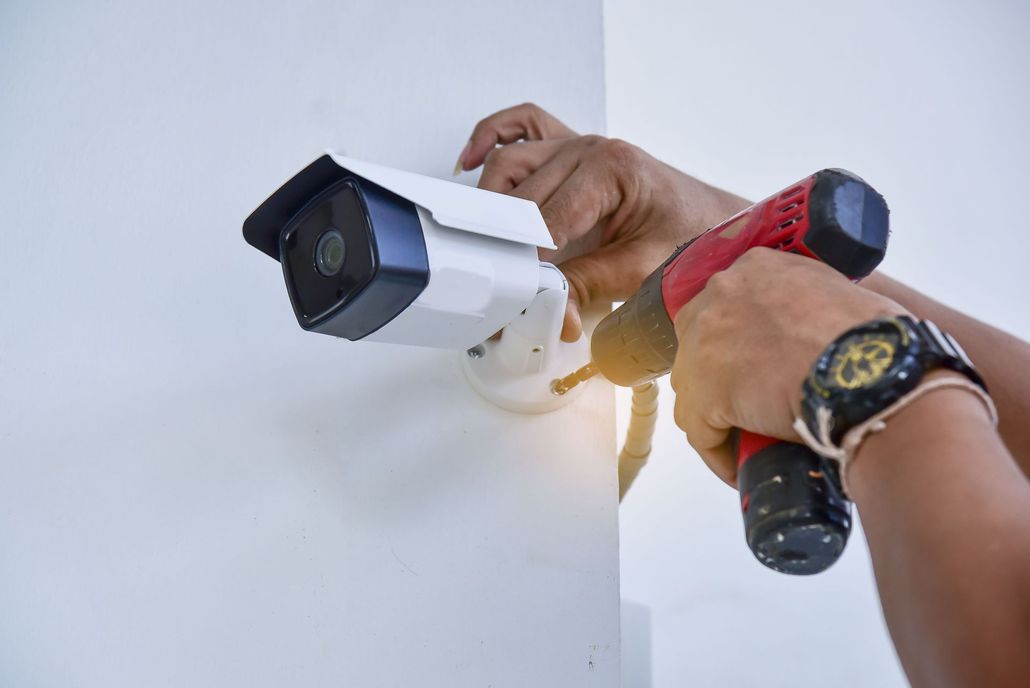 Hands using a power drill to install a white security camera on a white wall.