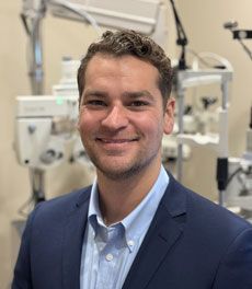 Man in blue blazer smiles in an optometrist's office, with eye examination equipment visible.