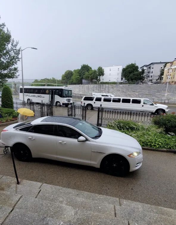 White Jaguar sedan with black wheels in front of a white limousine and bus, on a rainy street.