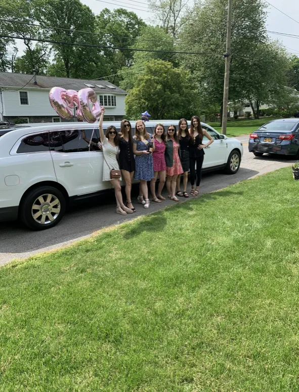 A group of women in dresses pose by a white limousine, holding pink heart balloons.