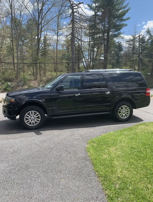 Black Ford Expedition SUV parked on a paved driveway next to green grass, trees in the background.