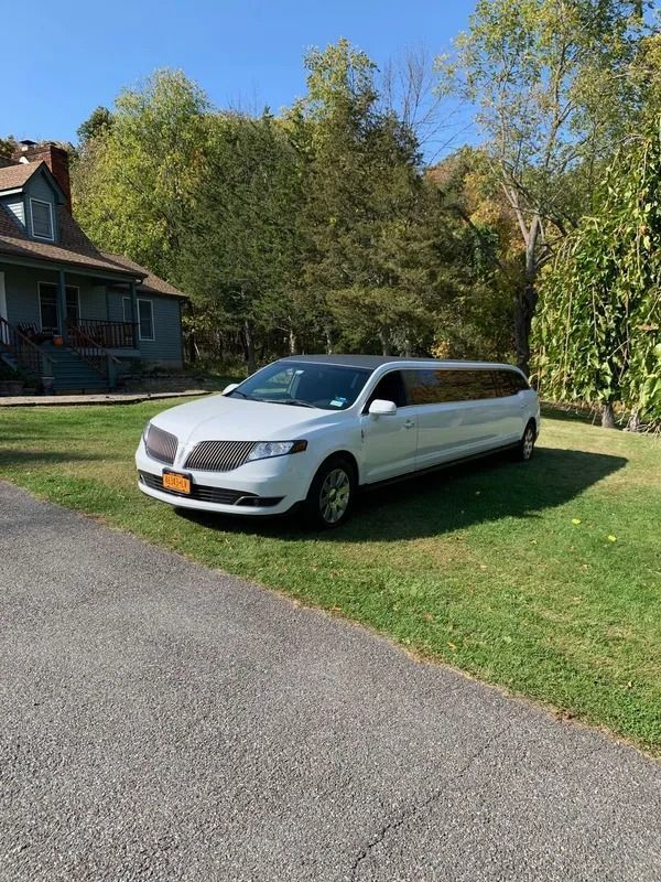 White Lincoln limousine parked on a lawn, next to a paved driveway and a blue house, under a sunny sky.