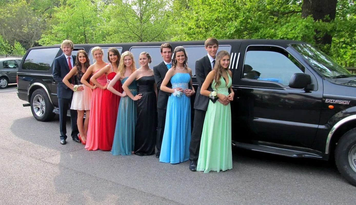 A group of teens in formal wear pose by a black limousine outdoors.