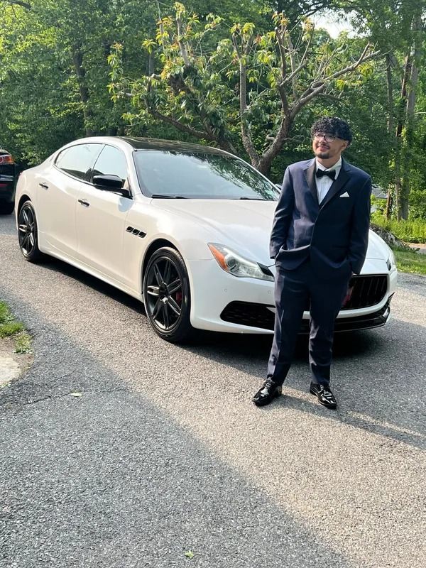 Man in a navy suit stands next to a white Maserati sedan on a paved road, trees in the background.