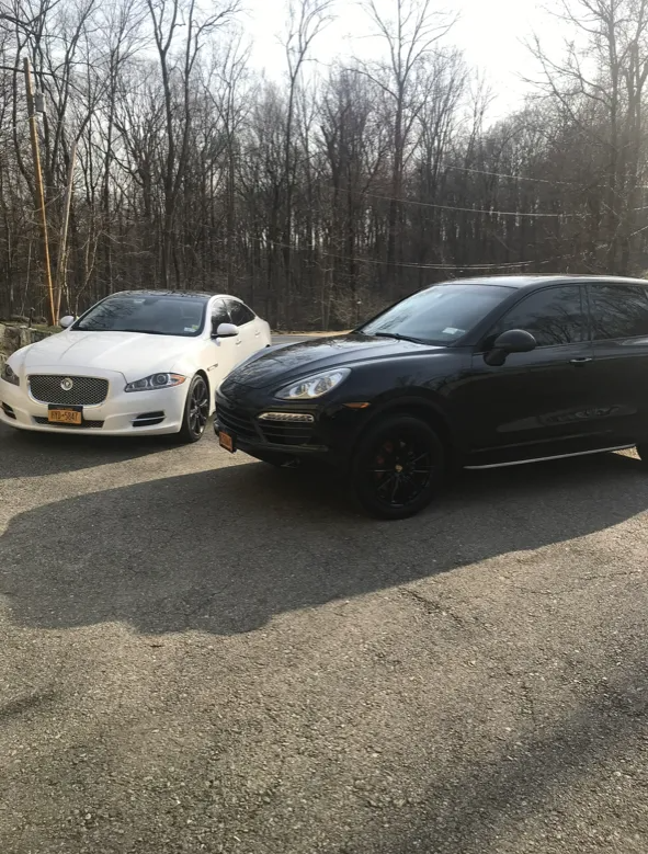 White Jaguar sedan and black Porsche SUV parked on pavement, trees in background.
