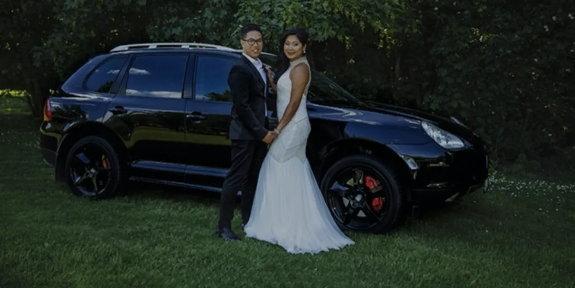 Newlyweds stand beside a black SUV on a grassy area, holding hands and smiling.