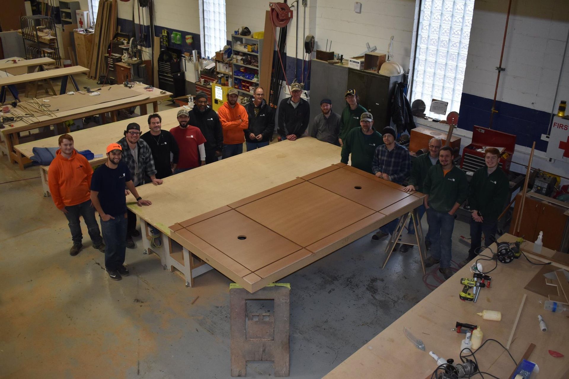 A large group of people stands in a woodshop around a newly constructed, flat wooden platform.