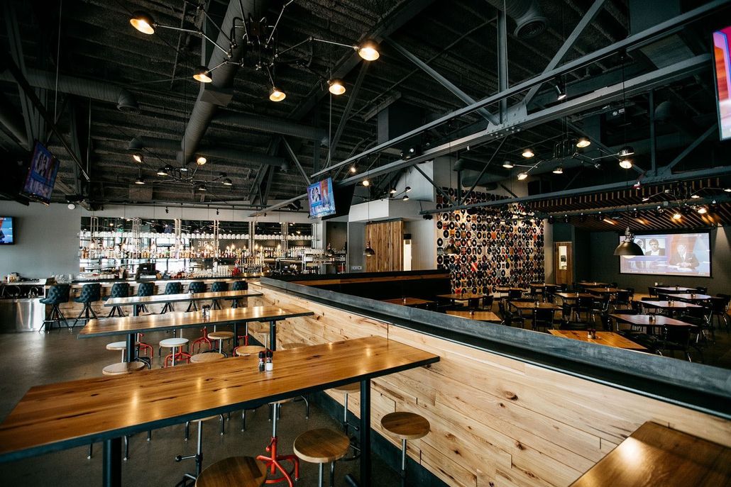 A modern, industrial-style restaurant interior with wooden tables, stools, an open ceiling, and a feature wall.