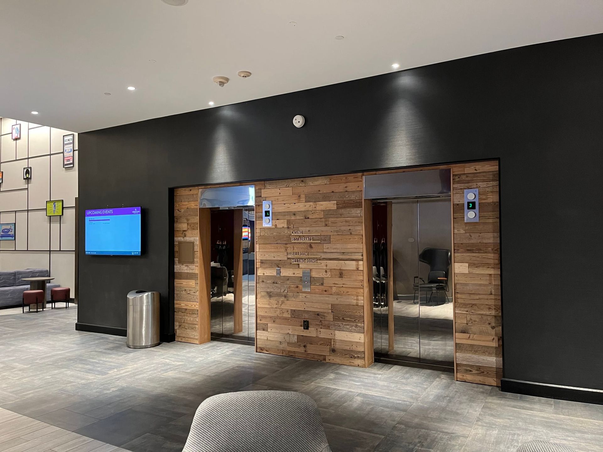 Elevator entrance with wood paneling. Two mirrored doors, dark gray wall with a TV screen and trash bin.