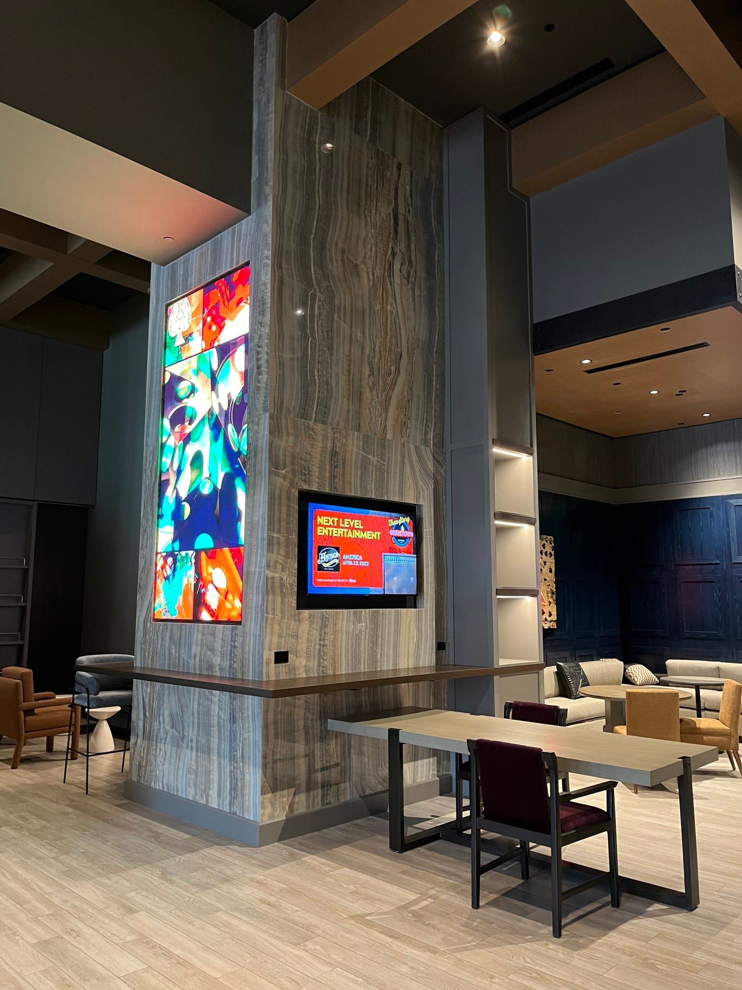 Lobby with digital displays on a pillar with wooden paneling. Tables, chairs, and seating area in the background.