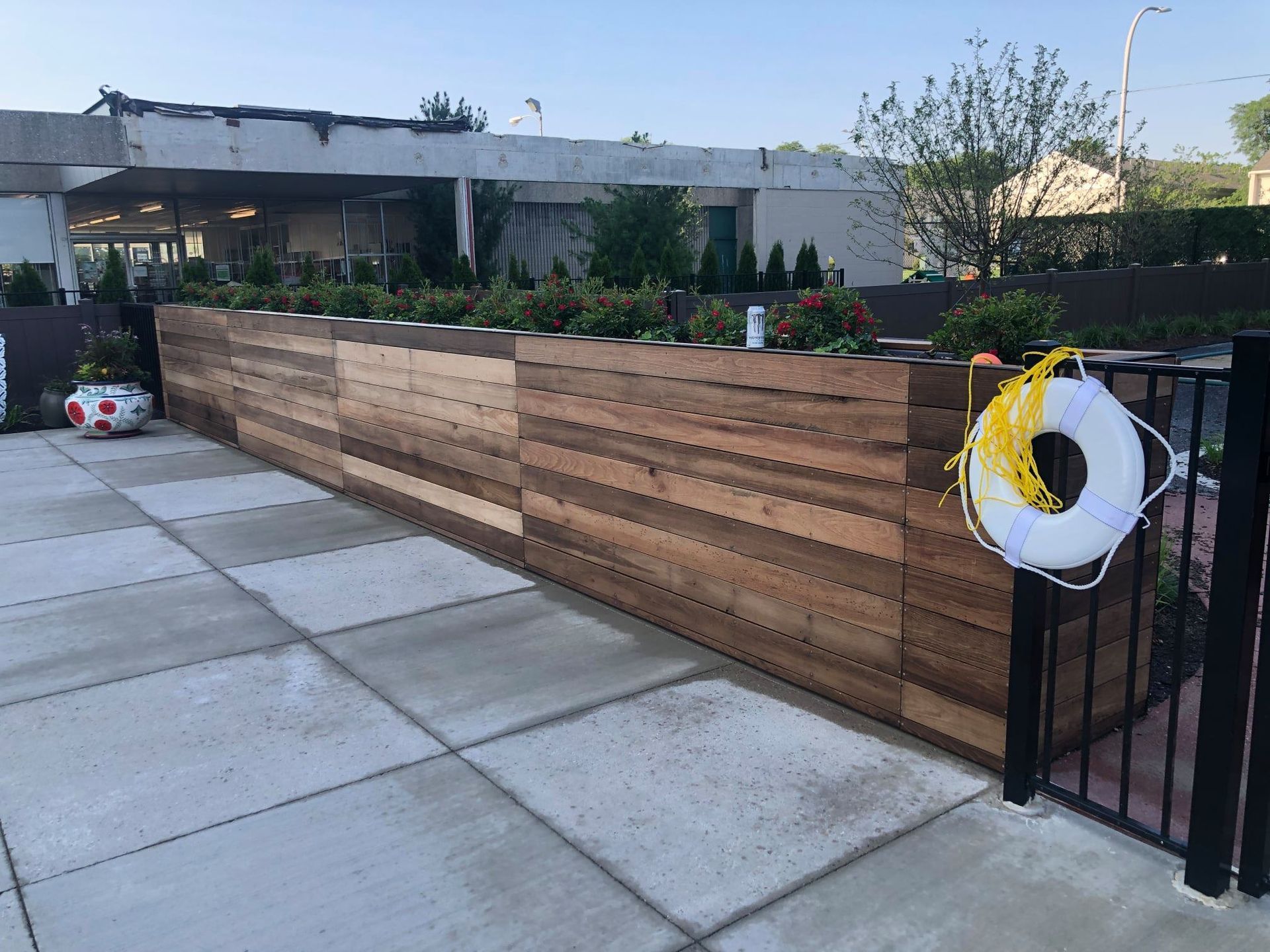 Wooden planter boxes with plants along a paved area, a building in the background, a life preserver and soccer ball nearby.