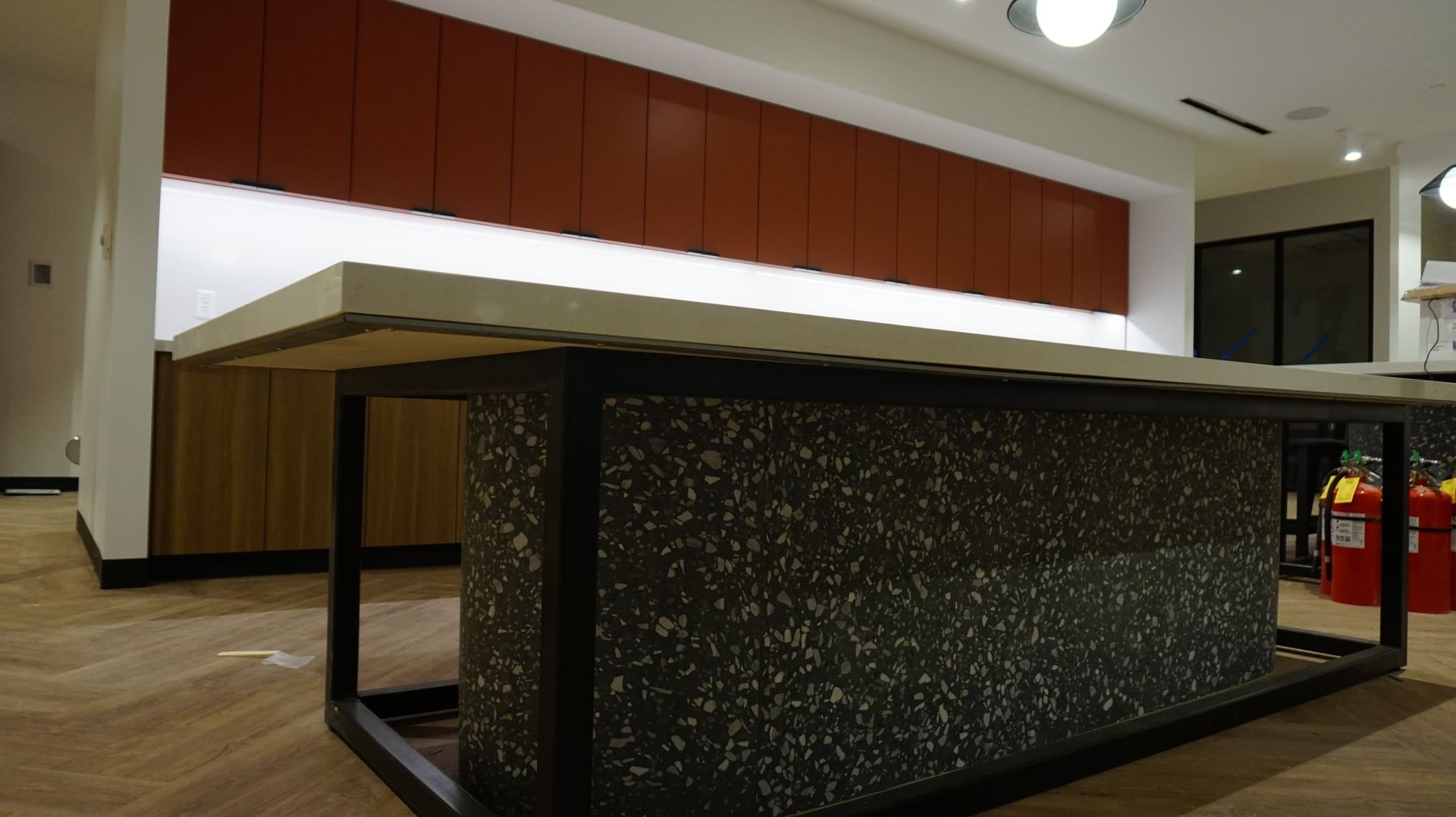 Large kitchen island with speckled gray facade and black metal frame. Red cabinets are in the background.