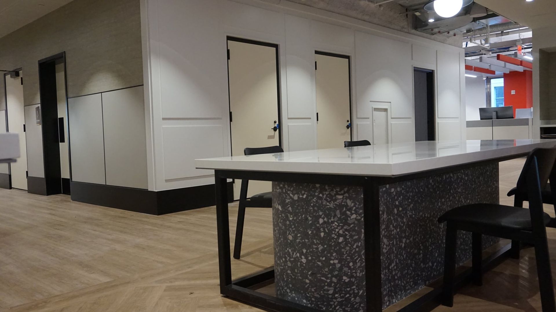 An office area with white walls, doors, and a table with black chairs. Wooden floor.
