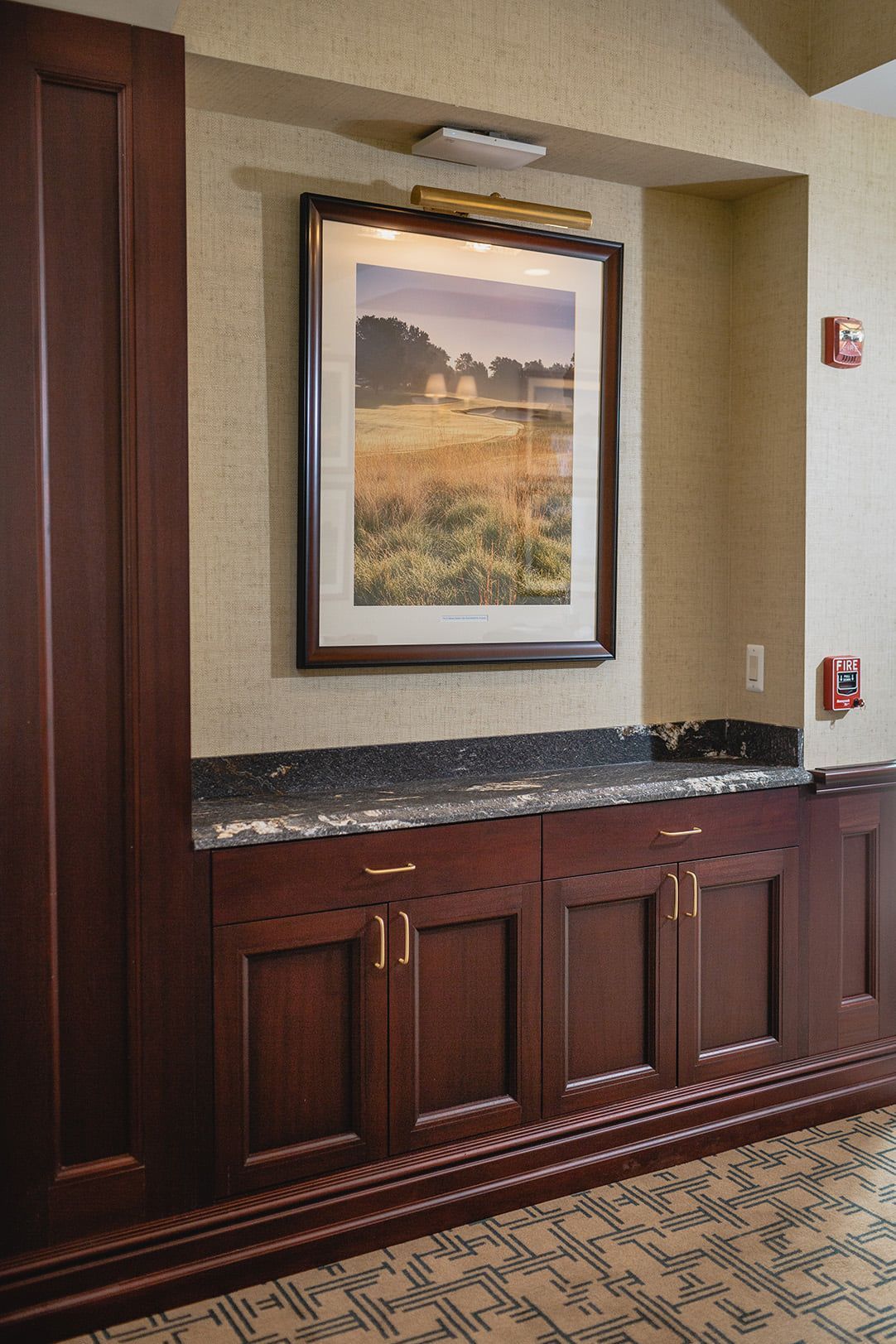 Built-in cabinetry with artwork above, lit by a lamp. Dark wood cabinets with a granite countertop.