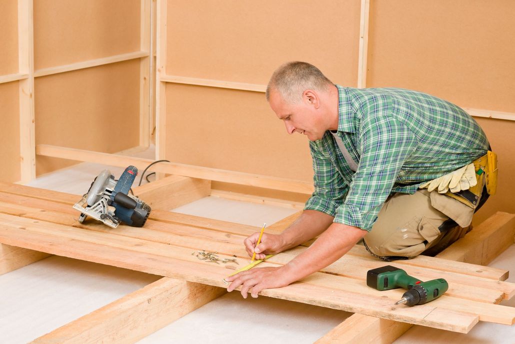 A person in a plaid shirt kneels on wooden floor joists, using a pencil and square to mark a board for cutting.