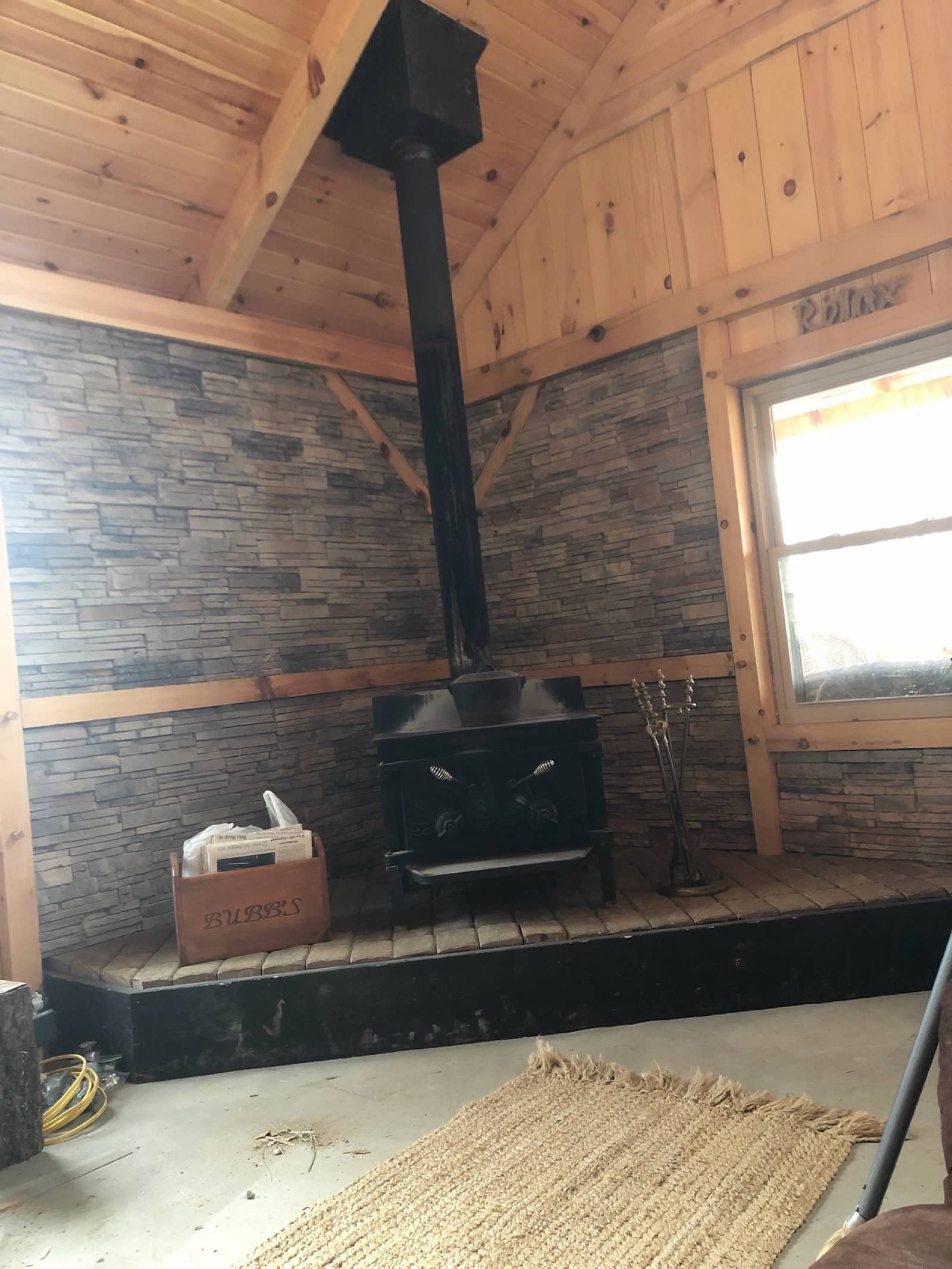Wood-paneled cabin interior with a black wood-burning stove and chimney. Stone-like wall covering and a small window.