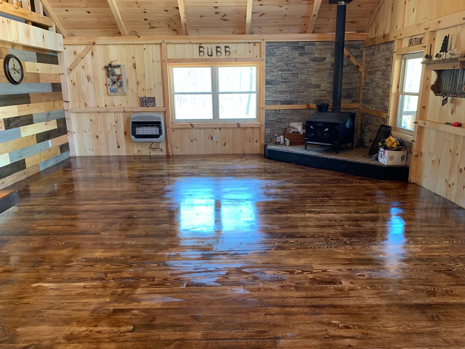 Wooden cabin interior with polished wood floor, wood-burning stove, and windows.