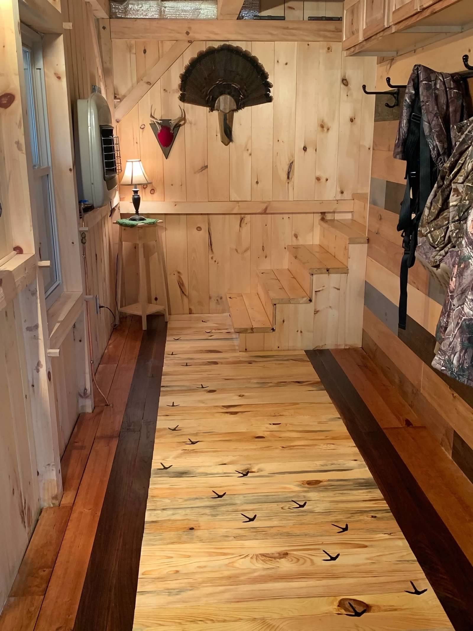 Interior of a tiny house with wood walls, floors, and stairs. Decorative antlers and camouflage clothing are visible.