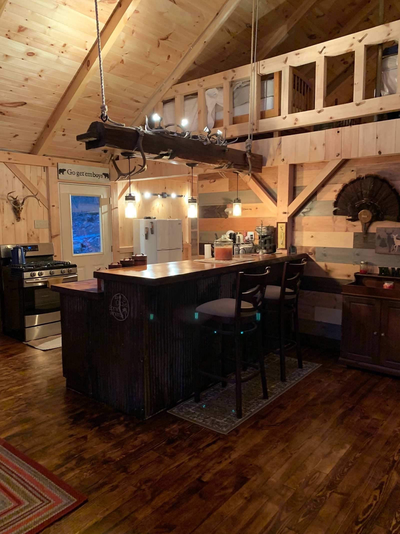Rustic kitchen with wooden bar, bar stools, and chandelier. Wooden beams, loft area, and hardwood floor.