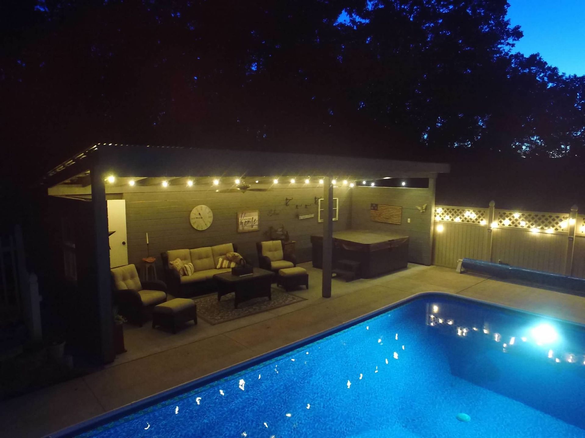 Nighttime view of a backyard patio with a pool. Lit with string lights, seating area, and hot tub.