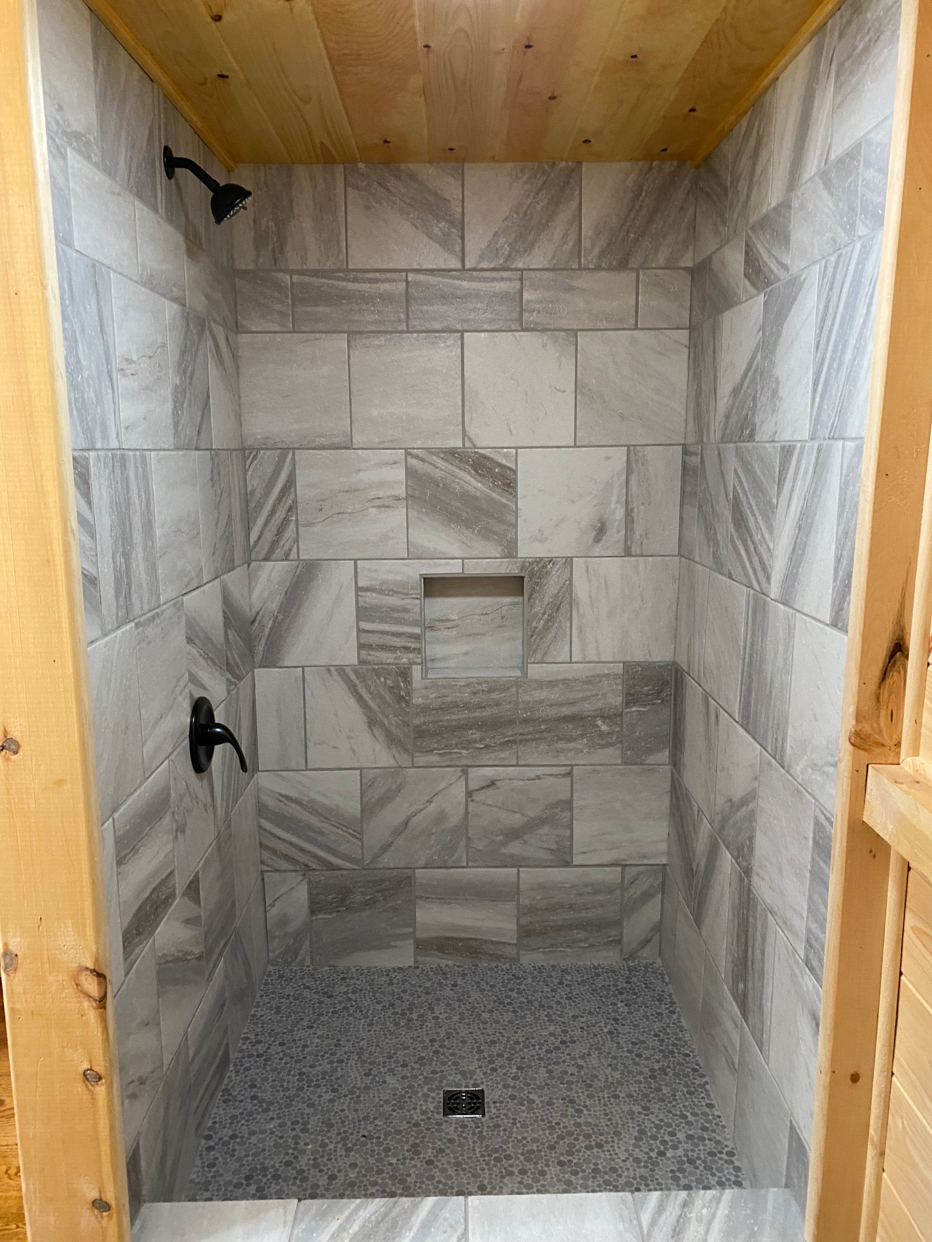 Tiled shower with gray stone-like tiles, a pebble floor, black fixtures, and wooden trim.