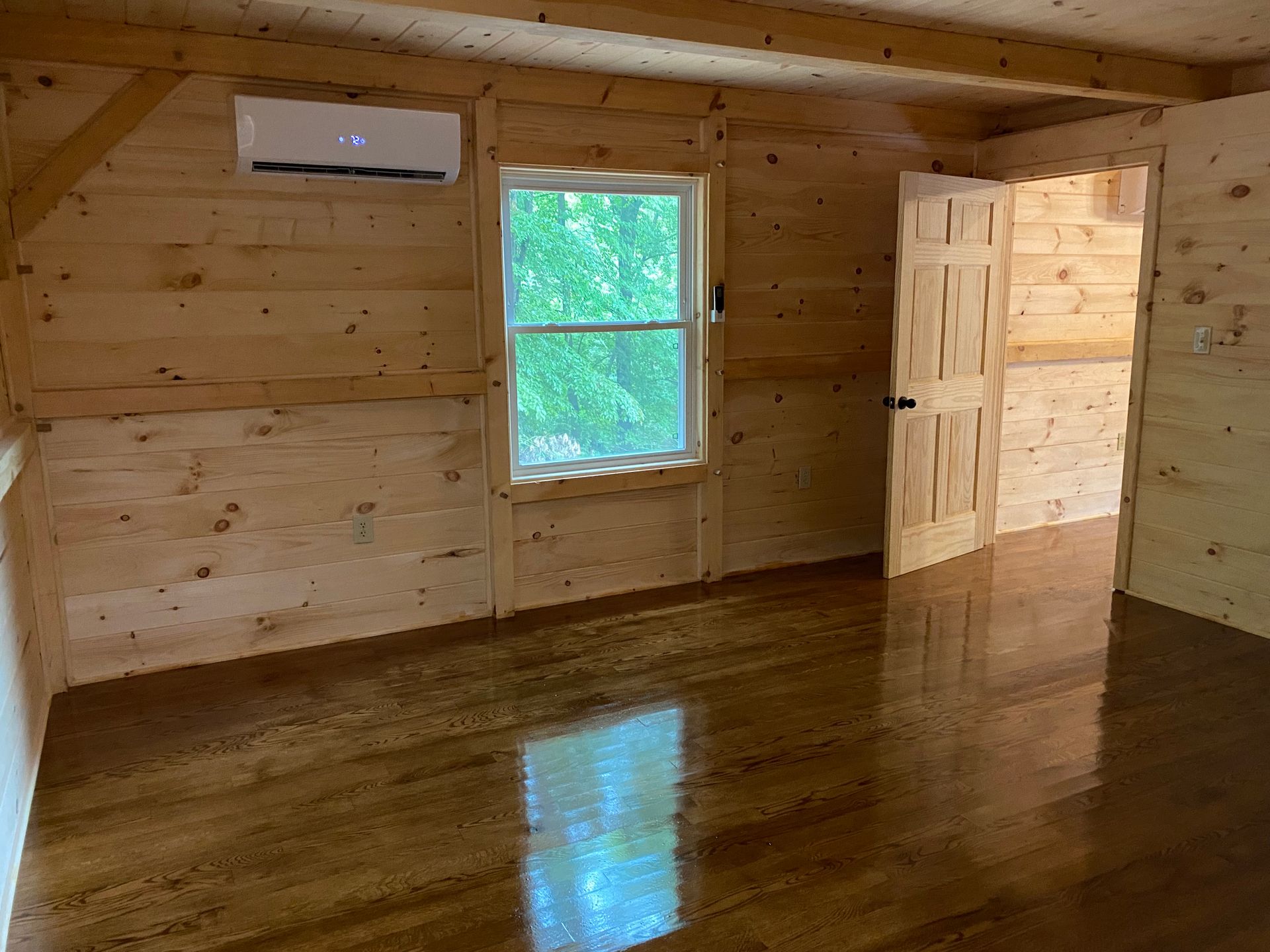 Interior of a room with wood paneling and glossy wooden floors. A window and door are visible. A/C unit on the wall.