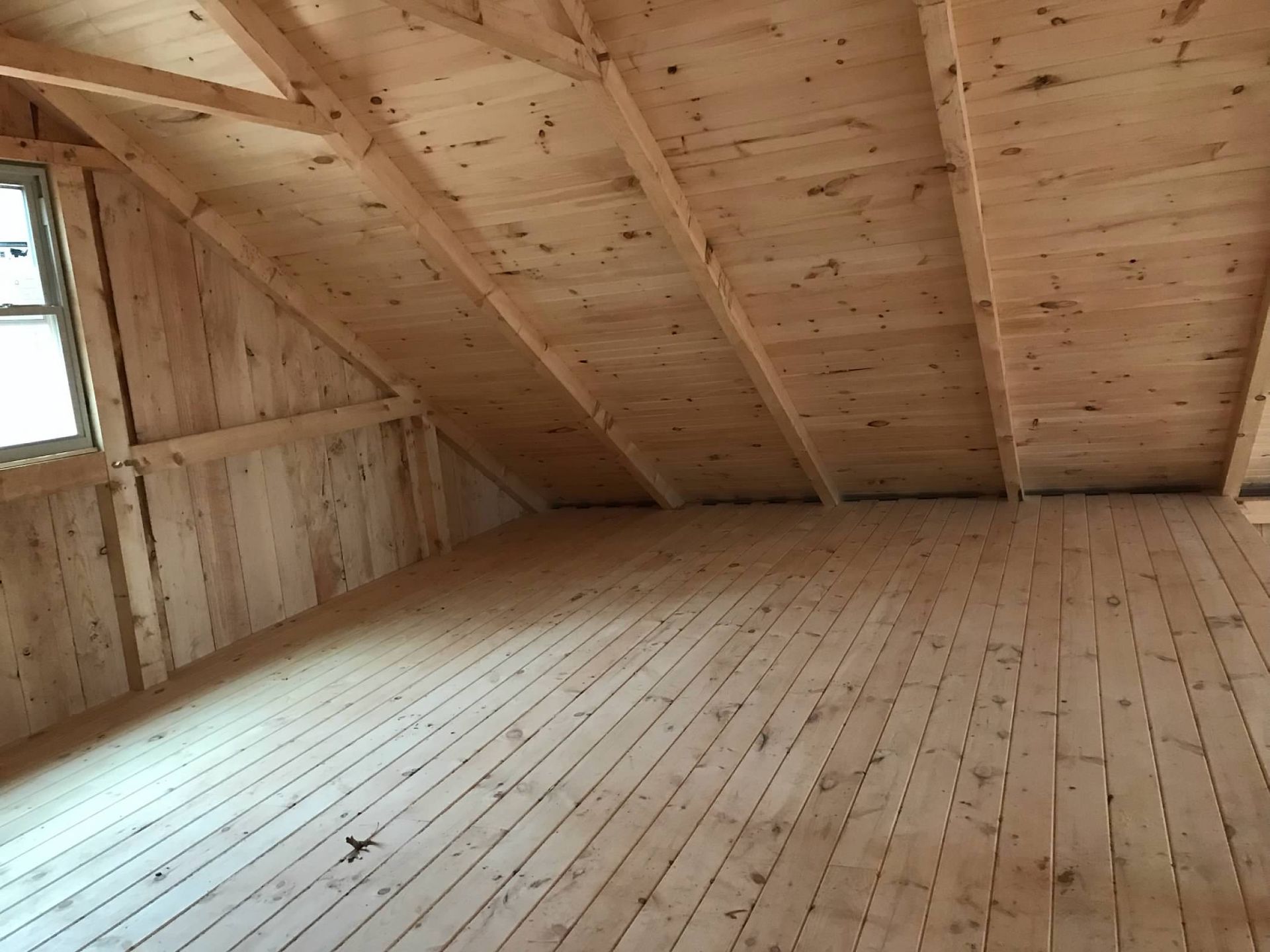 Interior view of unfinished wooden attic space with slanted ceiling, a window, and wood floor.