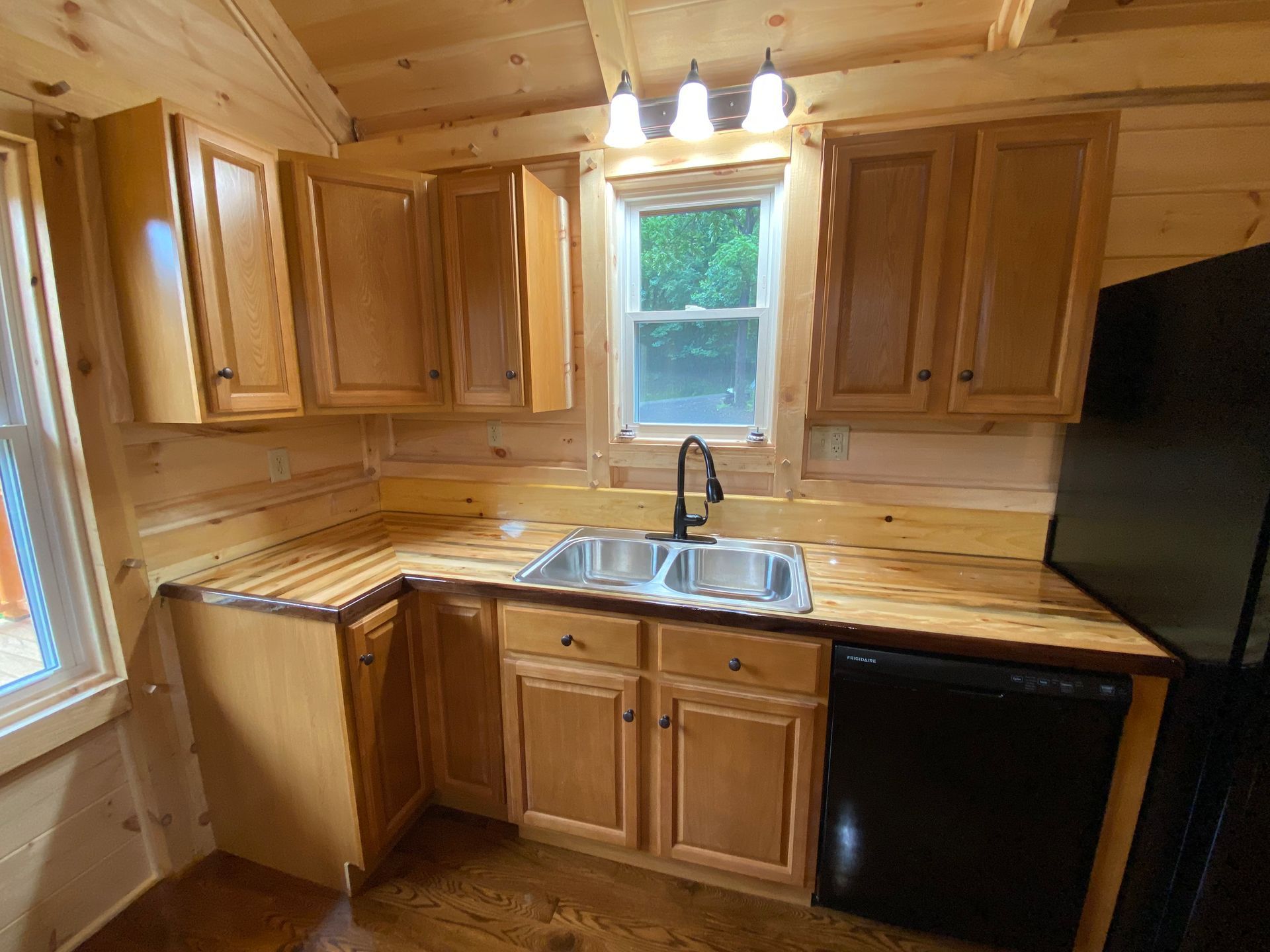 Cabin kitchen with light wood cabinets, double sink, black appliances, and small window.