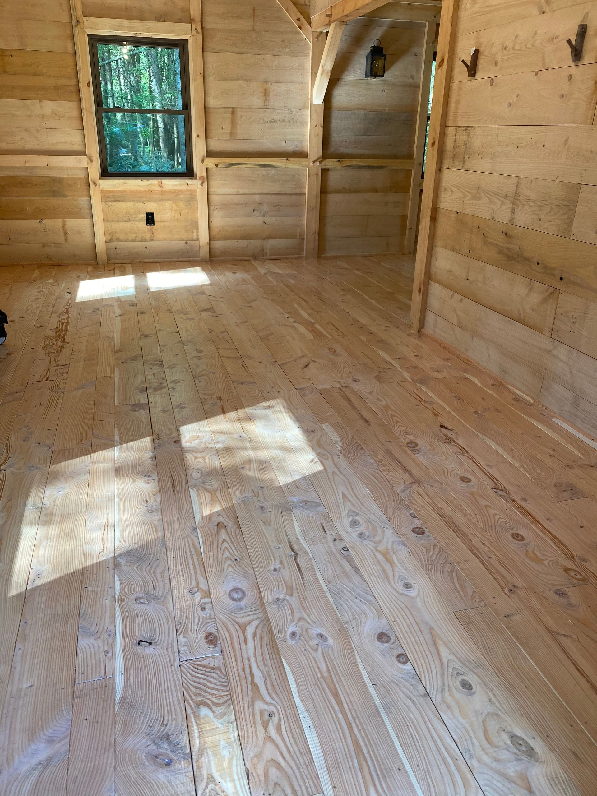 Wooden interior of a small cabin with sunlit floors and walls. A window and doorway are visible.