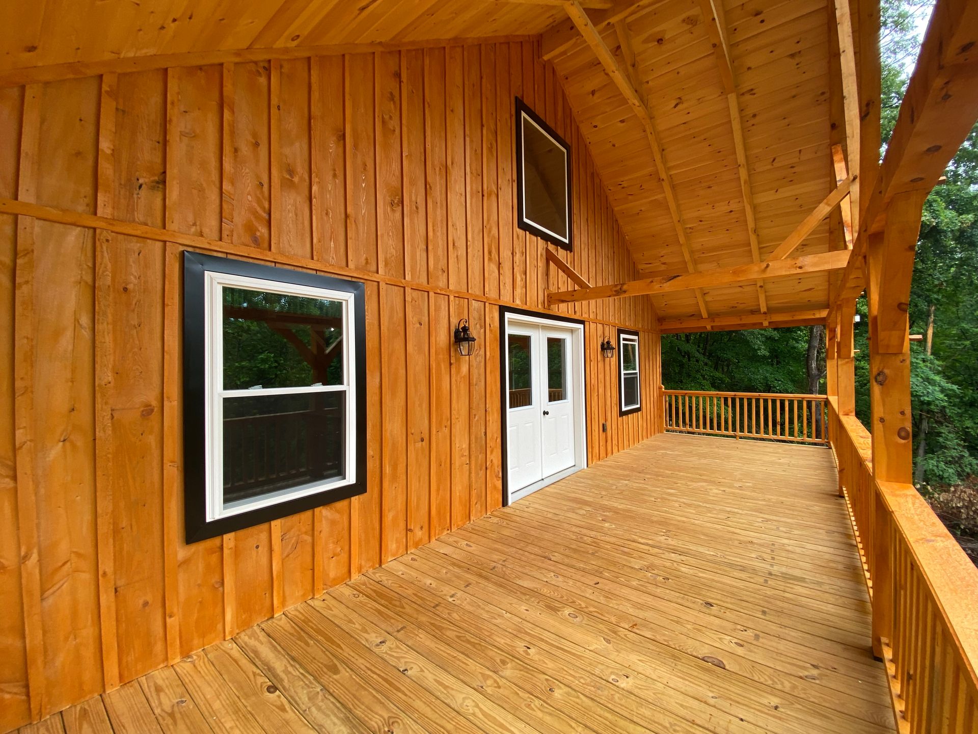 Wooden cabin with covered porch, windows, and white double doors. The building is surrounded by trees.
