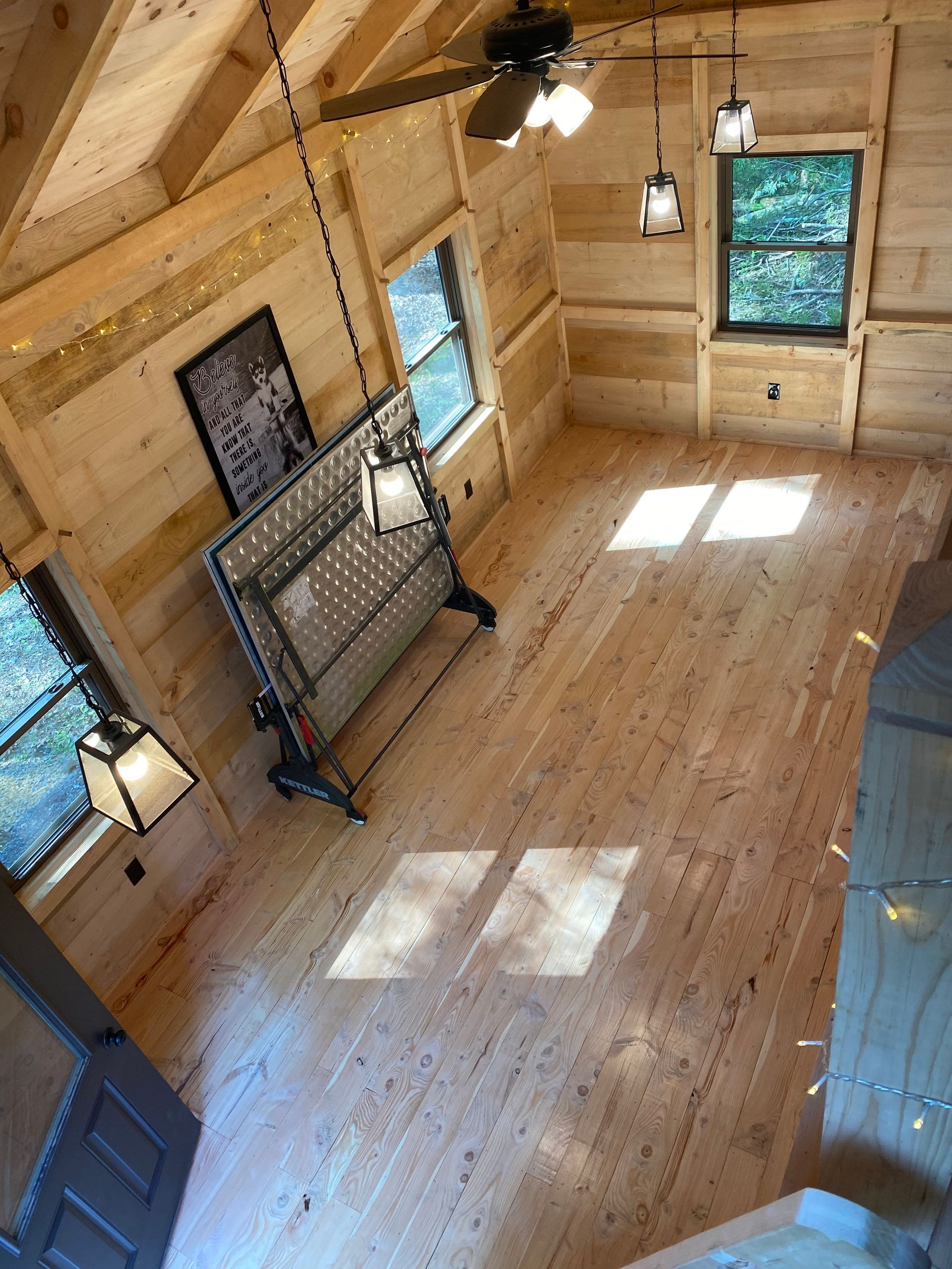 Interior of a small cabin with wooden walls, windows, bed, and lights. Sunlight streams in.