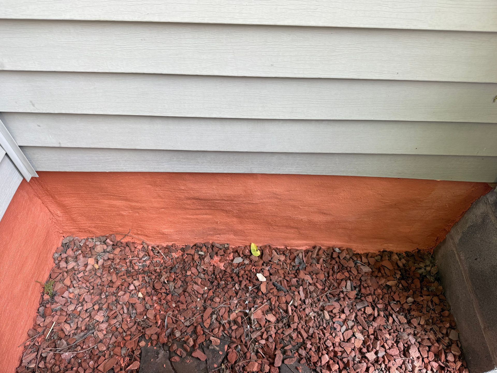 Reddish-brown mulch and ground cover beneath gray siding, a small sprout growing.