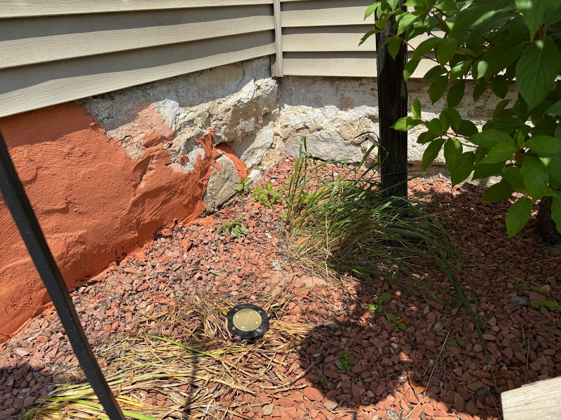 Red-brown stone foundation corner with damaged areas, red-brown mulch, small plants, and a dark tree trunk.