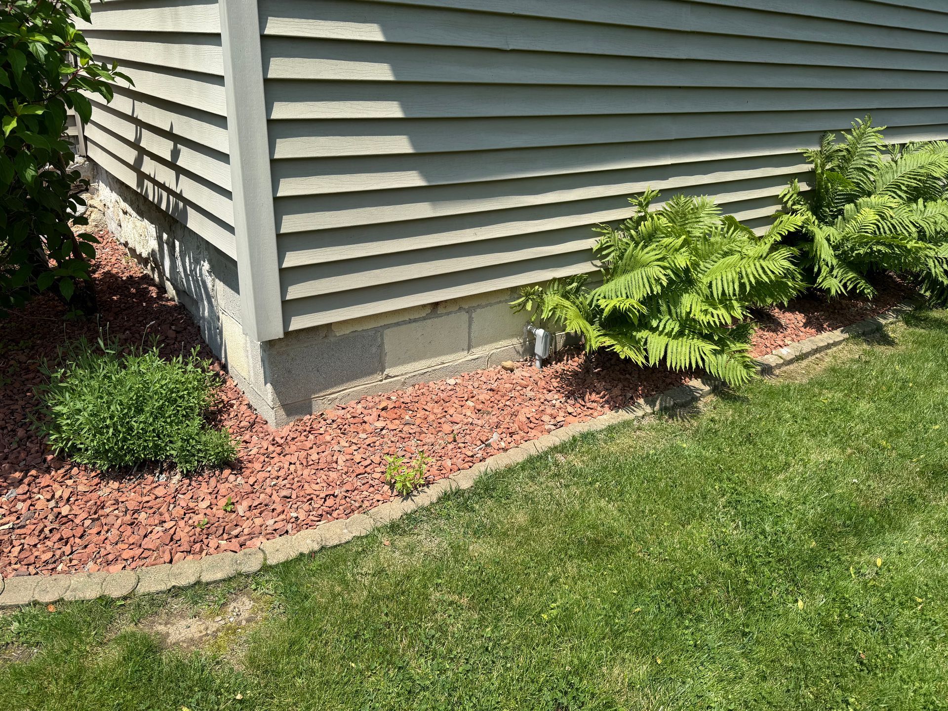 Green siding and corner of a building with red mulch and plants bordering the green grass.