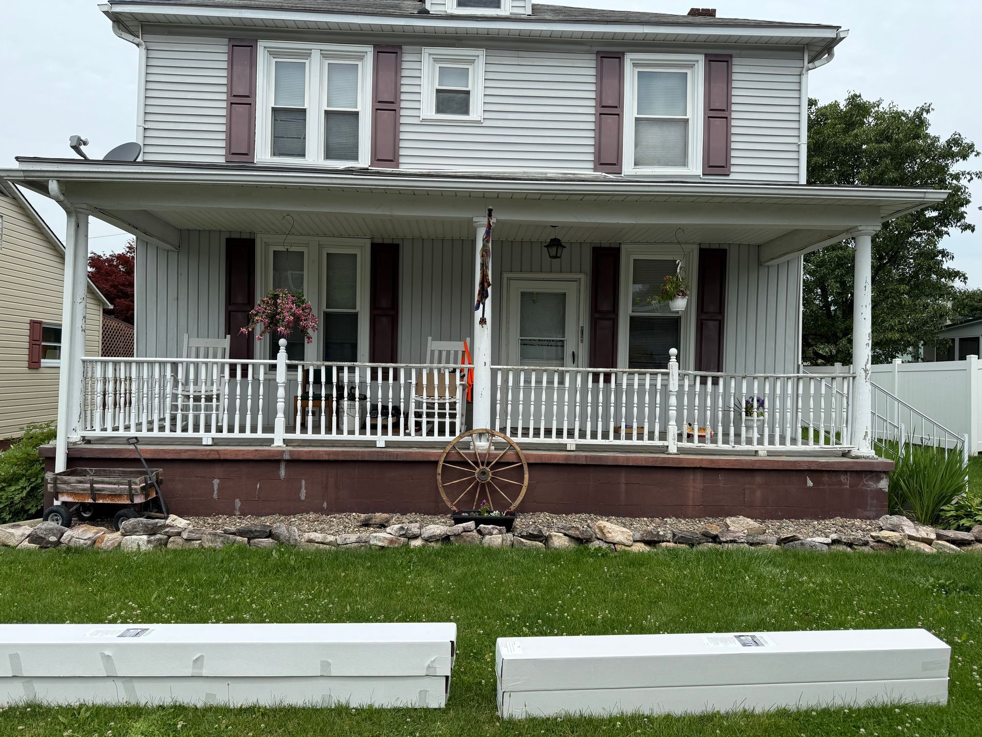 Two-story house with a porch and white picket fence. Red shutters. Green lawn with two white planks in the foreground.