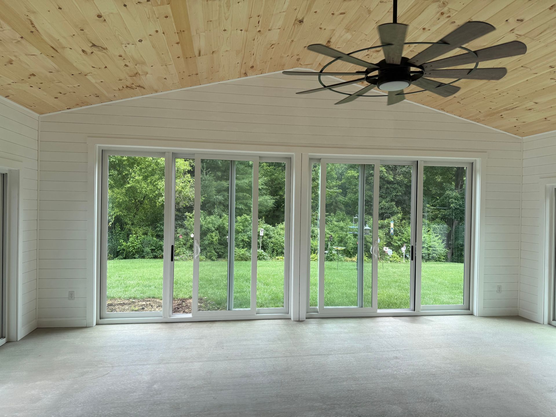 Bright sunroom with large glass doors overlooking a green yard, white walls, and a wood plank ceiling.
