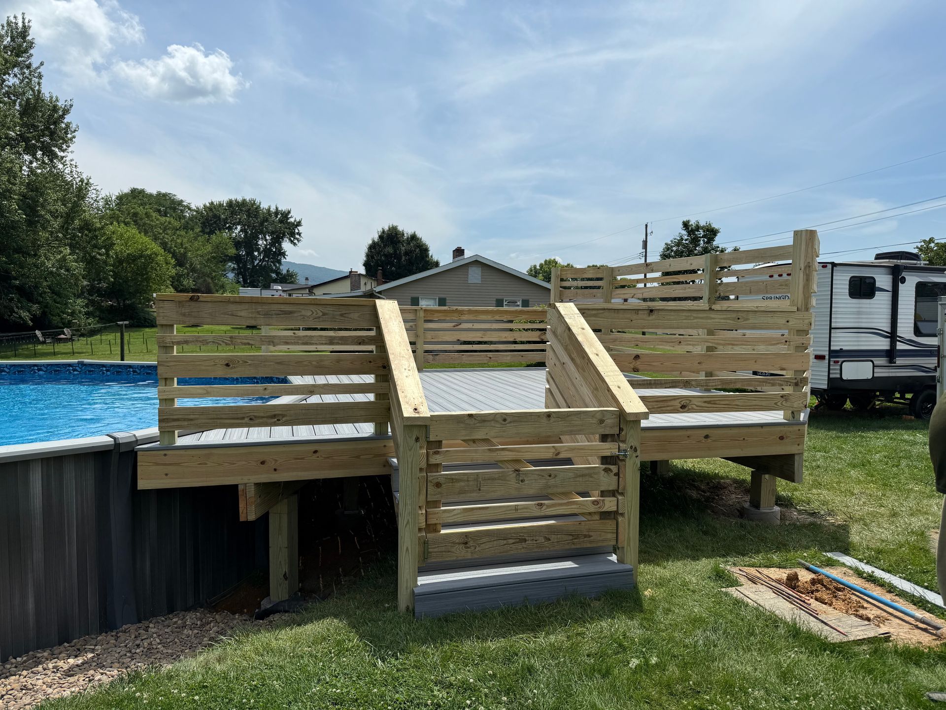 Wooden deck with steps next to a blue above-ground swimming pool on a grassy lawn under a partly cloudy sky.