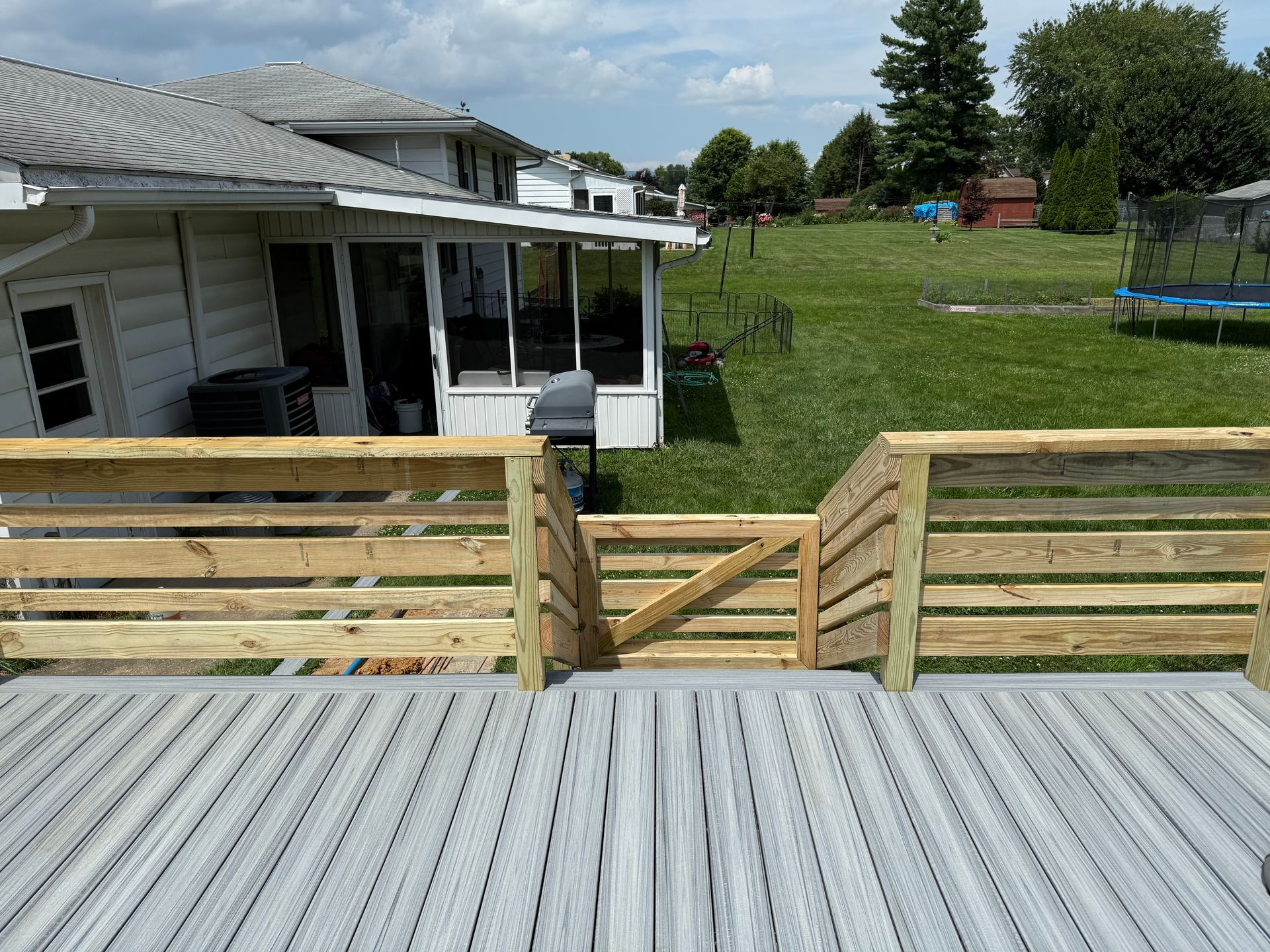 Wooden deck with light gray decking and light brown railings, overlooking a grassy backyard with houses in the background.