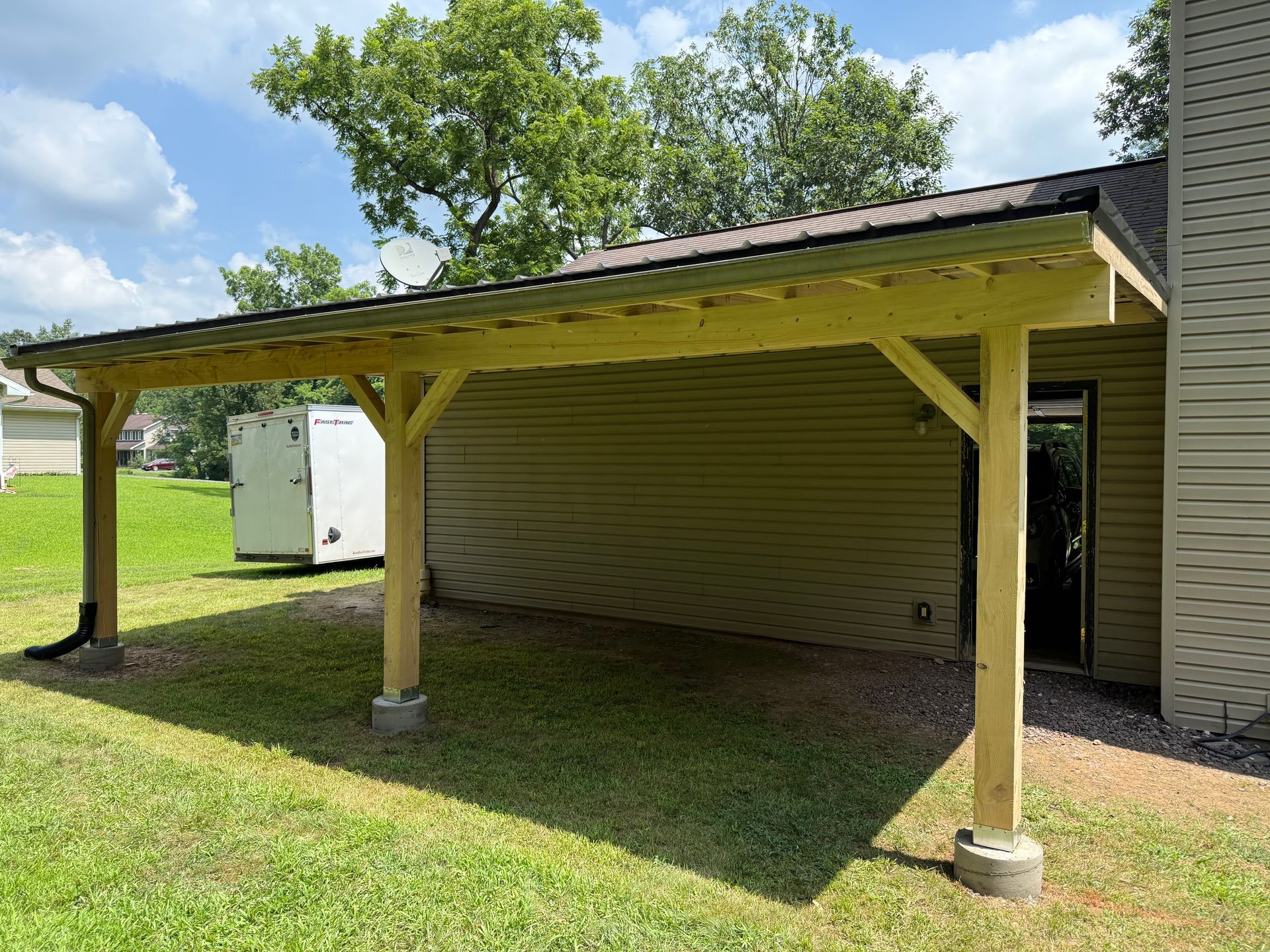A wooden carport with a metal roof attached to a beige house, on a green lawn.